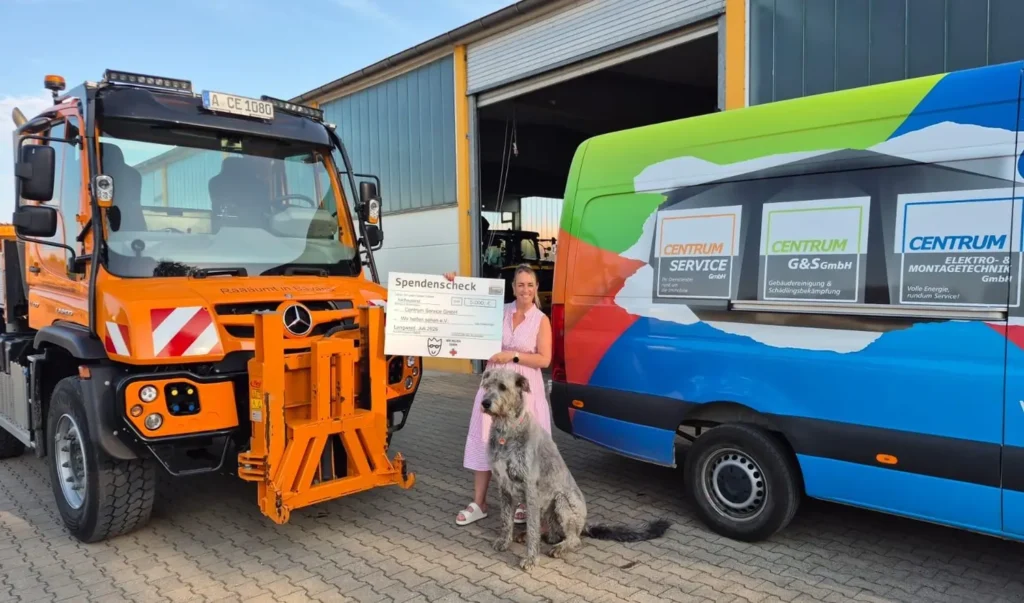 Frau steht zwischen zwei großen Autos und vor ihr sitzt ein Hund. Sie hält einen Spenden Banner in der Hand